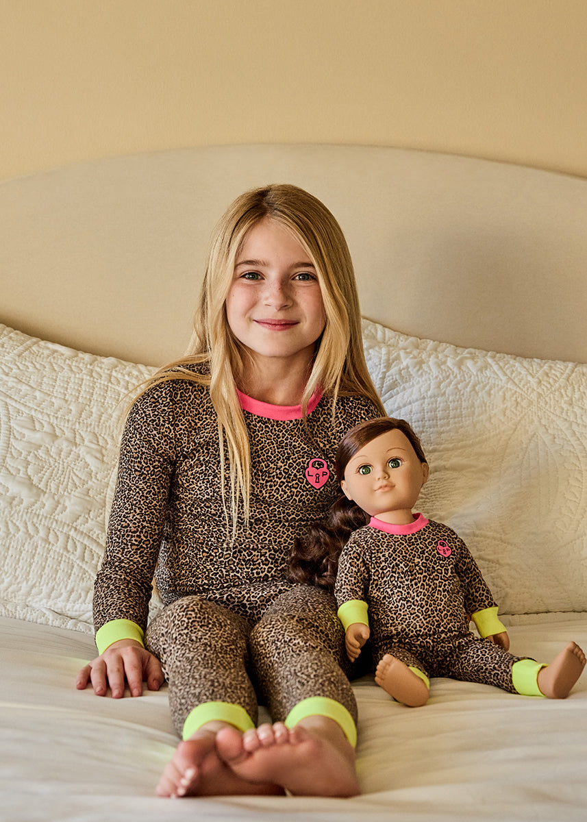 Young girl in brown pajamas with a matching doll on a bed.