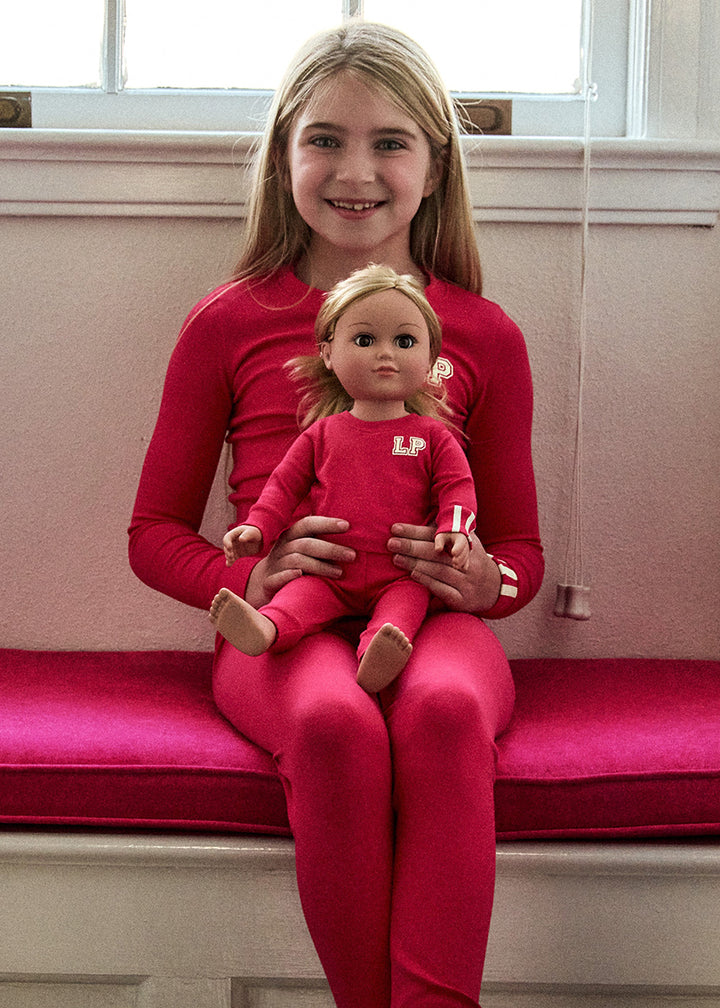 Young girl in red outfit holding a doll with a matching outfit on a pink cushioned bench.