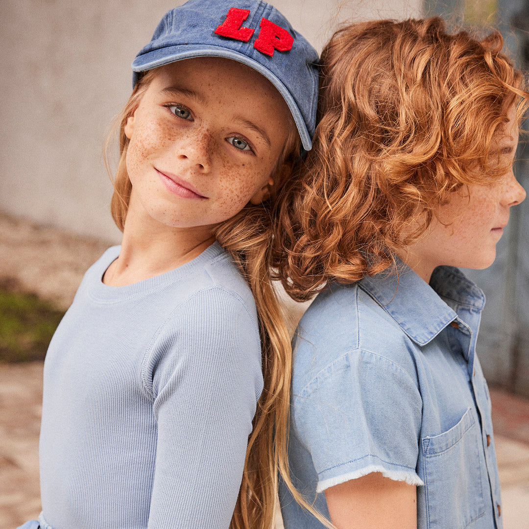 Close up of young girl and boy standing back to back outdoors wearing light denim tops and the girl is wearing a matching denim hat
