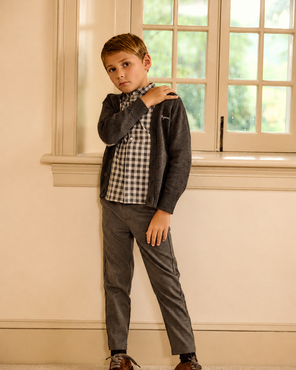 A young boy modeling a dark gray zip-up cardigan over a black and white gingham shirt, paired with gray trousers and brown leather shoes, standing in a room with cream walls and a window.