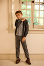 A young boy modeling a dark gray zip-up cardigan over a black and white gingham shirt, paired with gray trousers and brown leather shoes, standing in a room with cream walls and a window.