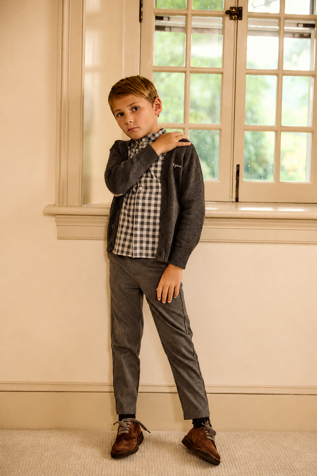 A young boy modeling a dark gray zip-up cardigan over a black and white gingham shirt, paired with gray trousers and brown leather shoes, standing in a room with cream walls and a window.
