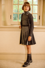 A young girl with a short brown bob stands indoors wearing a dark gray ribbed turtleneck sweater, matching pleated skirt, and dark gray knit socks and shoes. She is posed in front of a cream-colored wall and window.