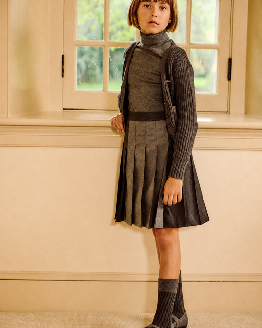A young girl with a short brown bob stands indoors wearing a dark gray ribbed turtleneck sweater, matching pleated skirt, and dark gray knit socks and shoes. She is posed in front of a cream-colored wall and window.