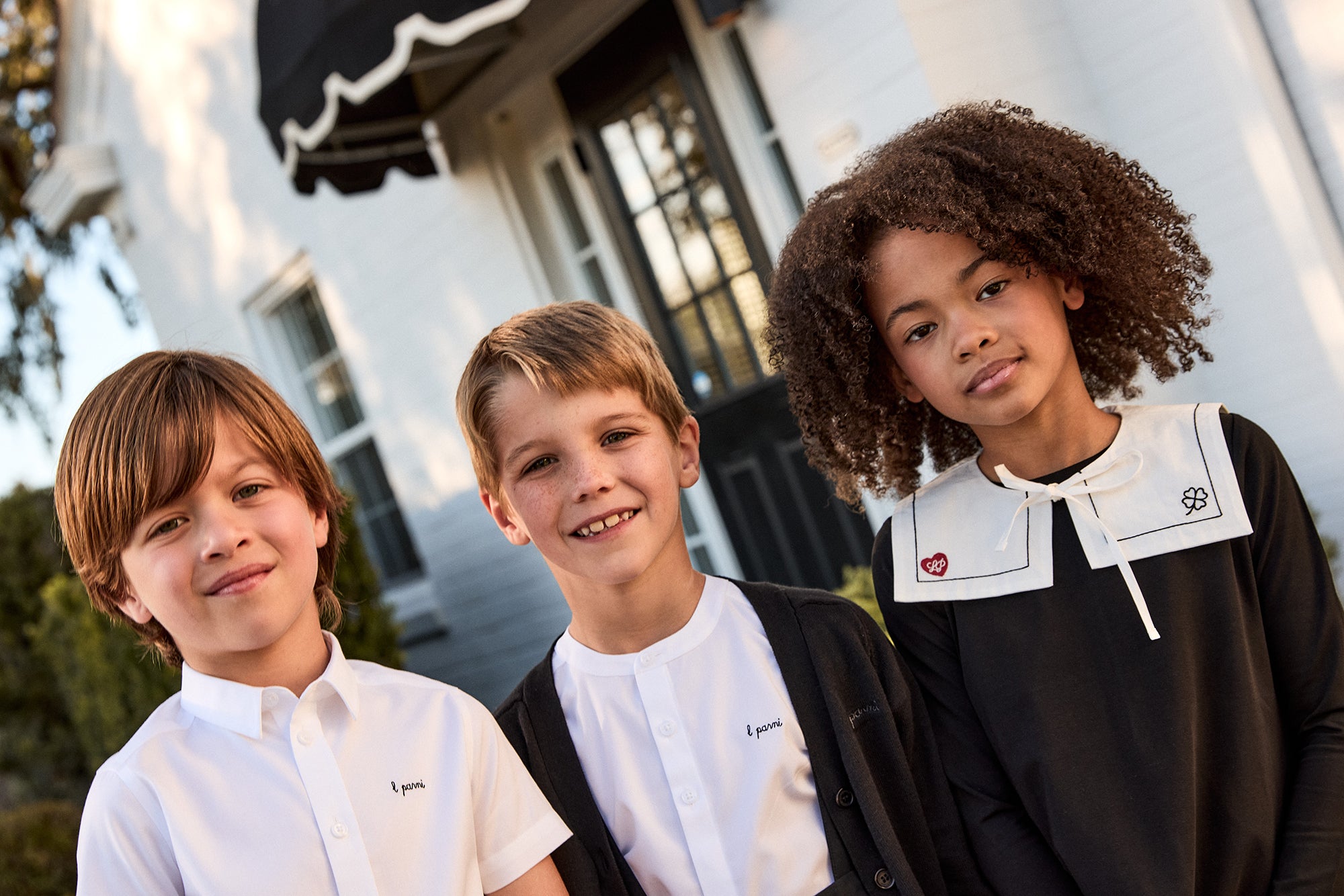 Three children standing together in front of a building with an awning.