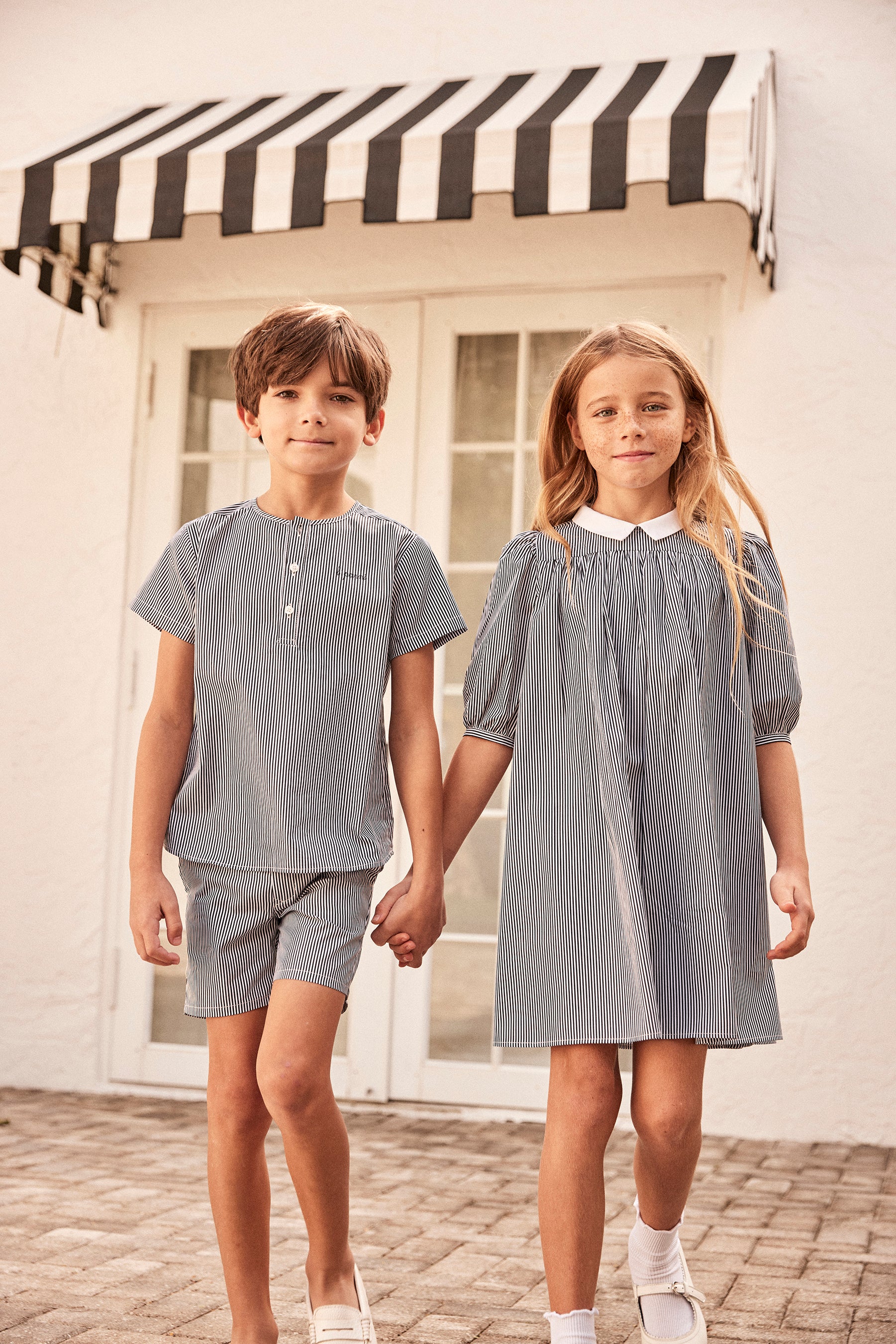 Young boy and girl wearing elegant matching outfits holding hands outside with a pretty home in the background.