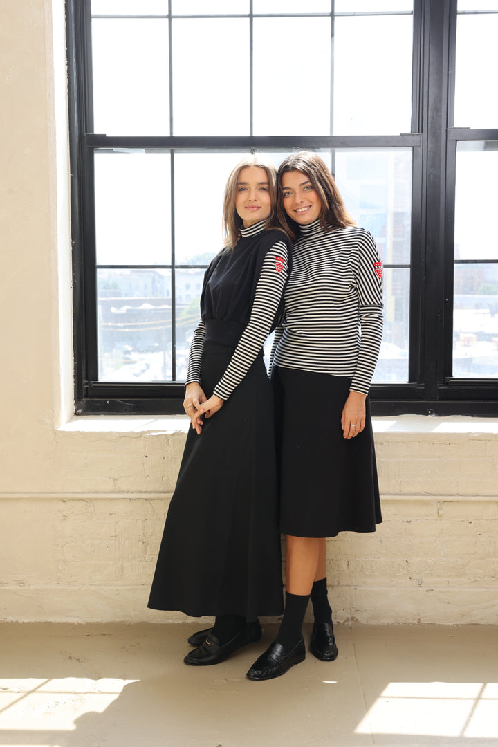Two women standing in a room with large windows, one wearing a black skirt and the other a striped shirt.