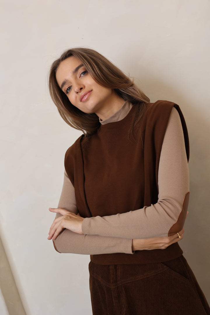 Woman wearing a brown sweater and beige shirt against a plain background
