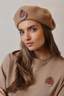 Woman wearing a brown beret with embroidered badges against a neutral background