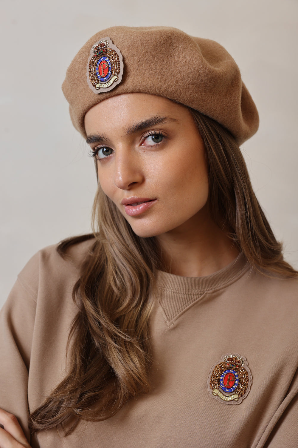 Woman wearing a brown beret with embroidered badges against a neutral background