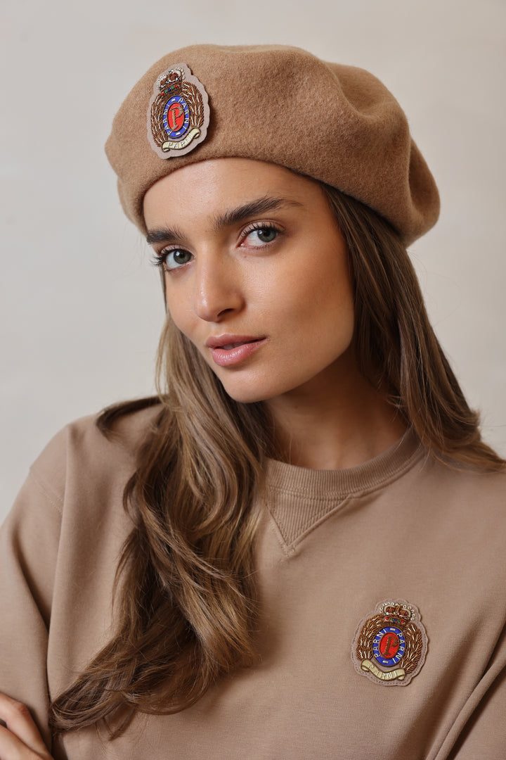 Woman wearing a brown beret with embroidered badges against a neutral background