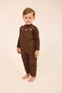 A toddler standing barefoot against a plain cream studio background, wearing a dark brown corduroy overall jumpsuit with long sleeves and silver-toned metal snap details at the chest.