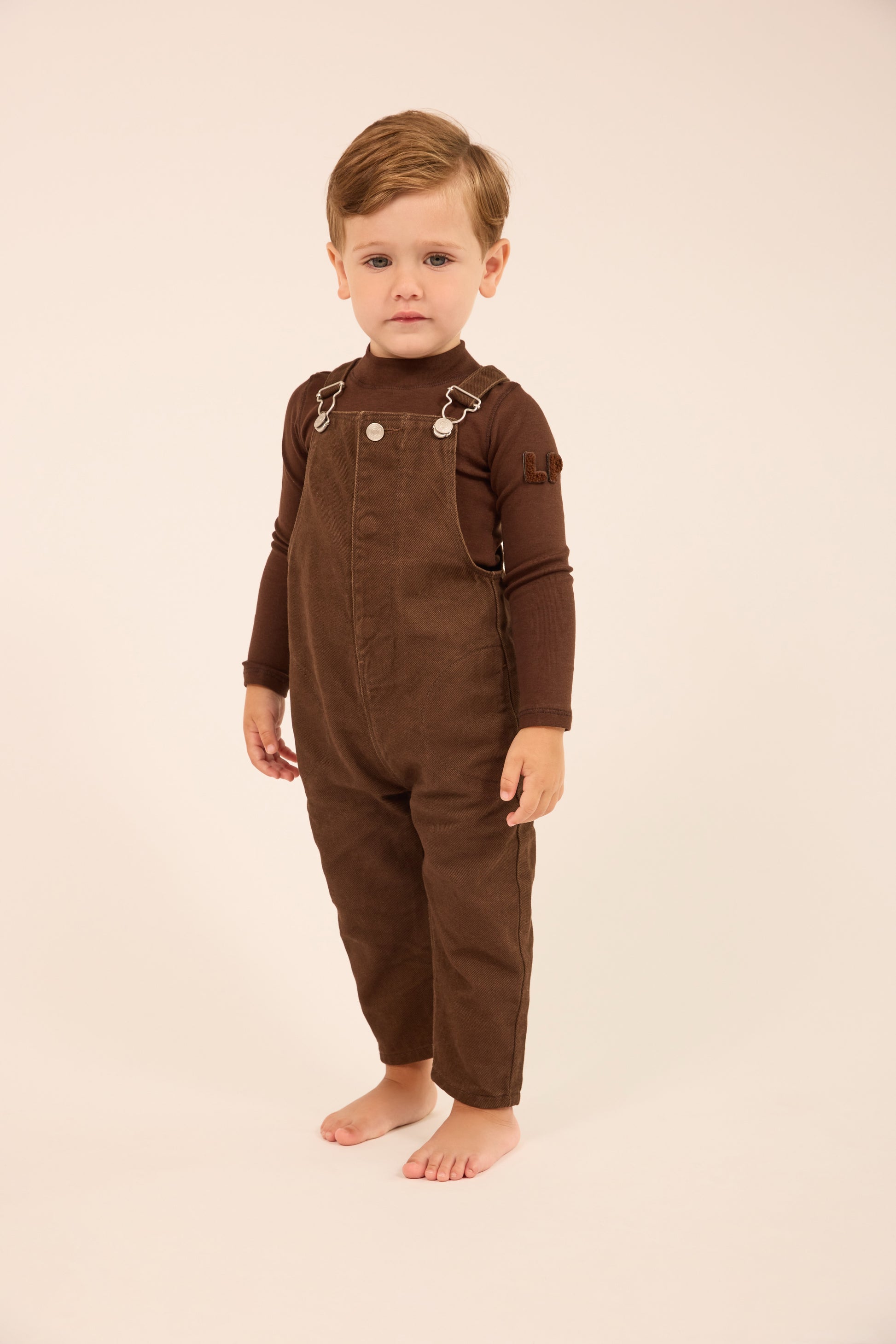 A toddler standing barefoot against a plain cream studio background, wearing a dark brown corduroy overall jumpsuit with long sleeves and silver-toned metal snap details at the chest.