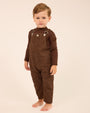 A toddler standing barefoot against a plain cream studio background, wearing a dark brown corduroy overall jumpsuit with long sleeves and silver-toned metal snap details at the chest.