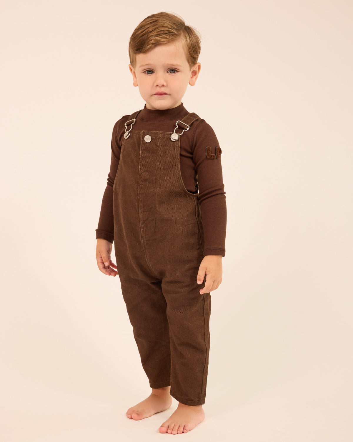 A toddler standing barefoot against a plain cream studio background, wearing a dark brown corduroy overall jumpsuit with long sleeves and silver-toned metal snap details at the chest.