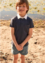 Young boy standing outdoors by a body of water, wearing a dark polo shirt with a white collar and light blue shorts.