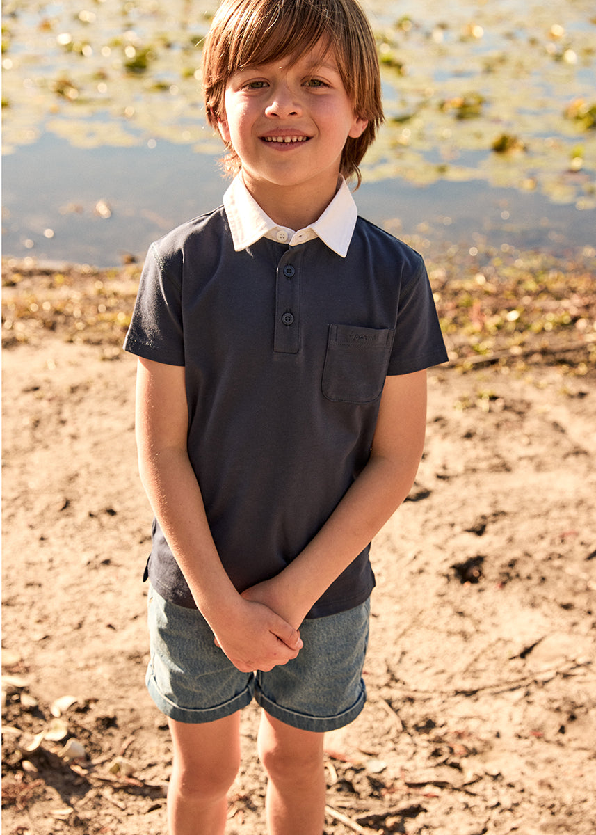 Young boy standing outdoors by a body of water, wearing a dark polo shirt with a white collar and light blue shorts.