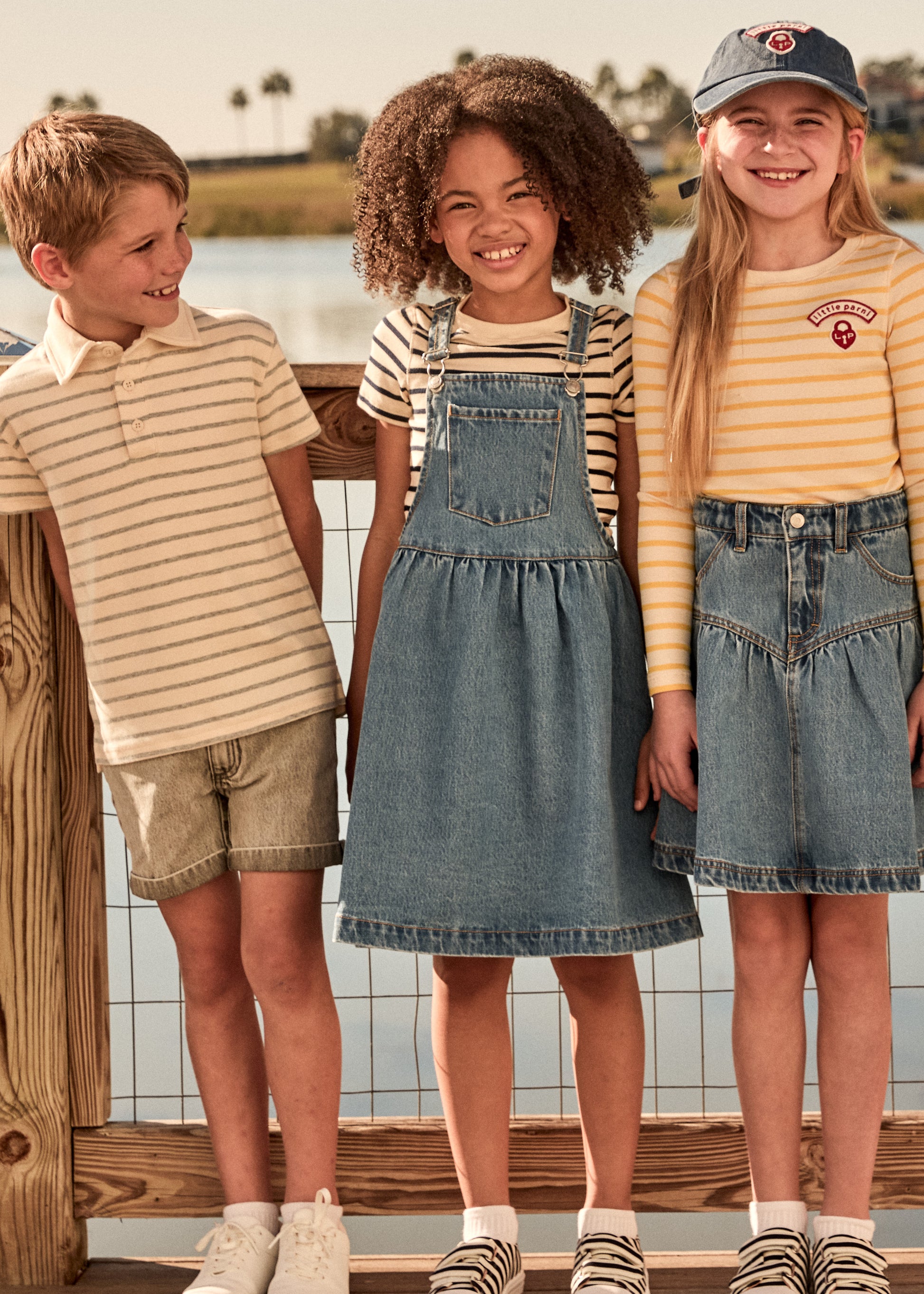 Three children standing by a lake, wearing matching denim skirts and striped shirts.