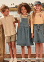 Three children standing by a lake, wearing matching denim skirts and striped shirts.