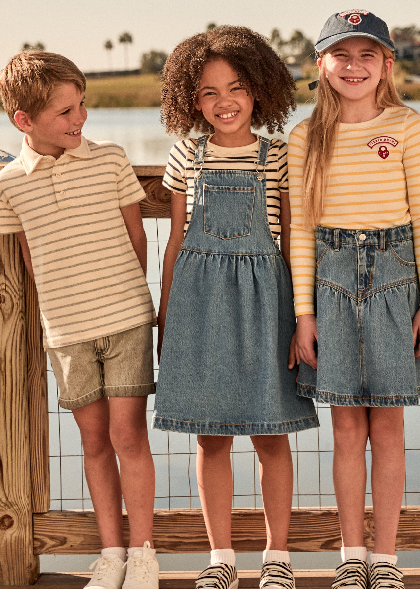 Three children standing by a lake, wearing matching denim skirts and striped shirts.