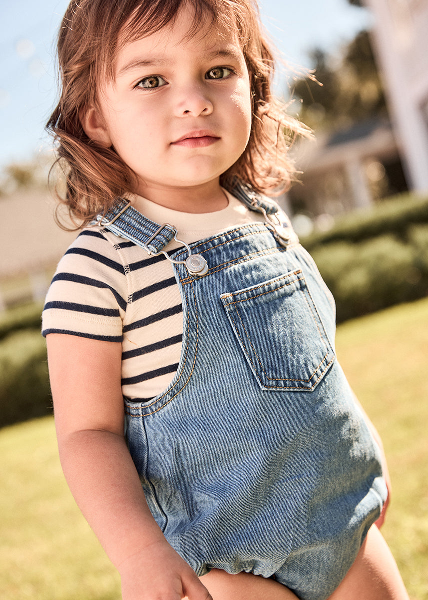 Child wearing a striped shirt and denim overalls sitting outdoors.