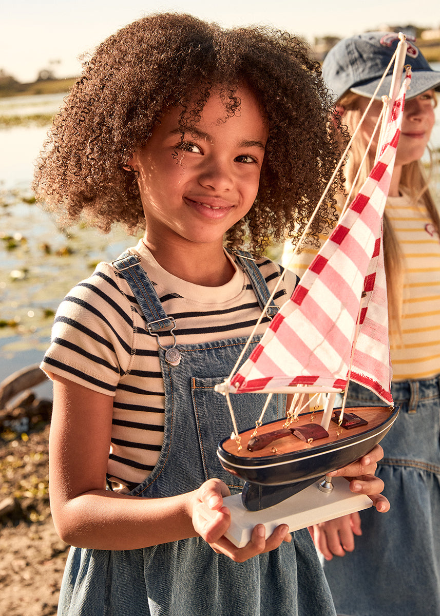 Child holding a model sailboat by a body of water with another person in the background.