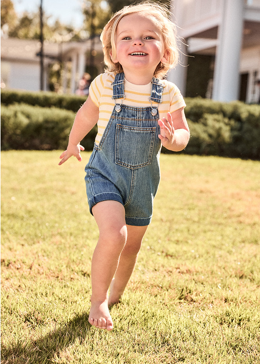 Child in denim overalls and yellow shirt standing on grass