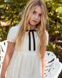 Young girl in a white dress with a black ribbon standing outdoors.