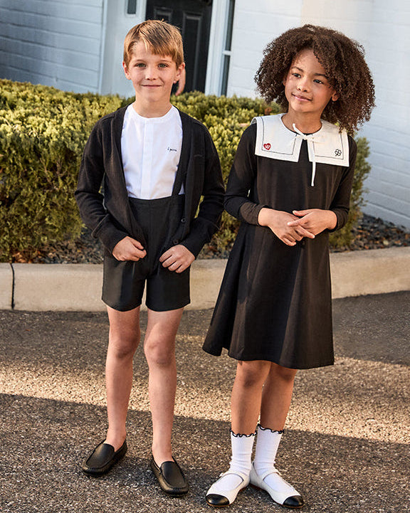Two children in formal attire standing in front of a white building with a black awning.