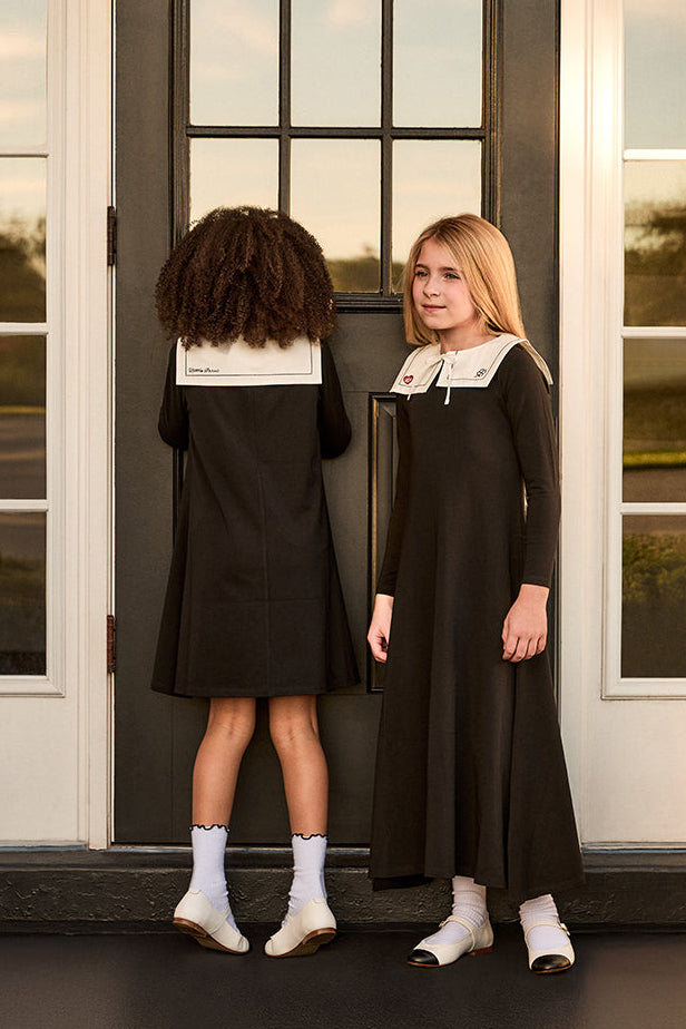 Two girls in black dresses with white collars standing in front of a glass door.