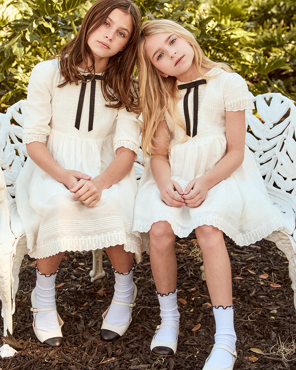 Two young girls in matching white dresses with black ribbons sitting on a bench outdoors.