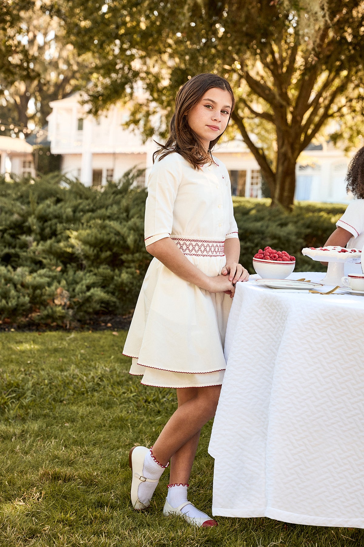 Young girl in a white dress standing outdoors near a table with a white tablecloth.