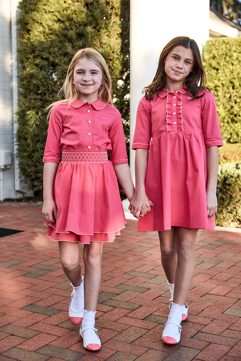 Two young girls wearing matching pink dresses standing on a brick path.