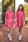 Two young girls wearing matching pink dresses standing on a brick path.