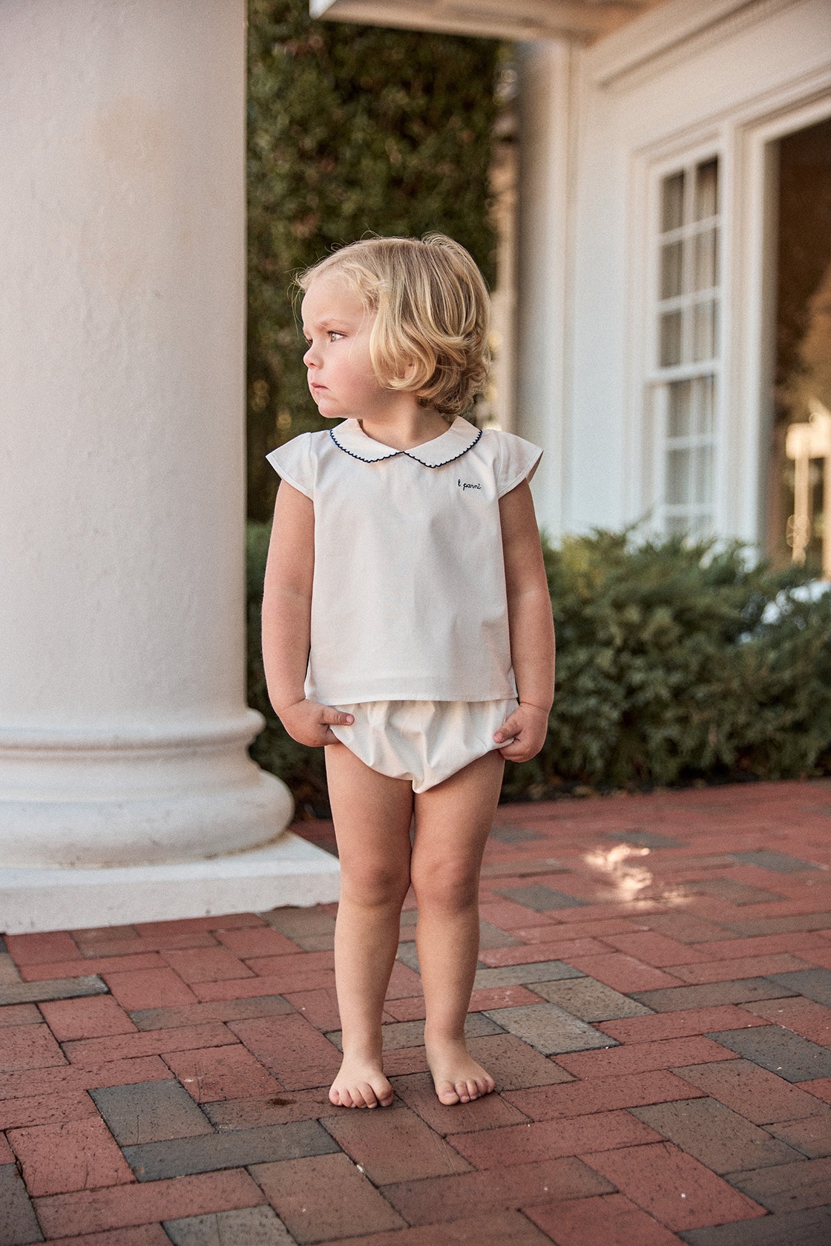 Young child standing on a brick patio in front of a white building with columns.