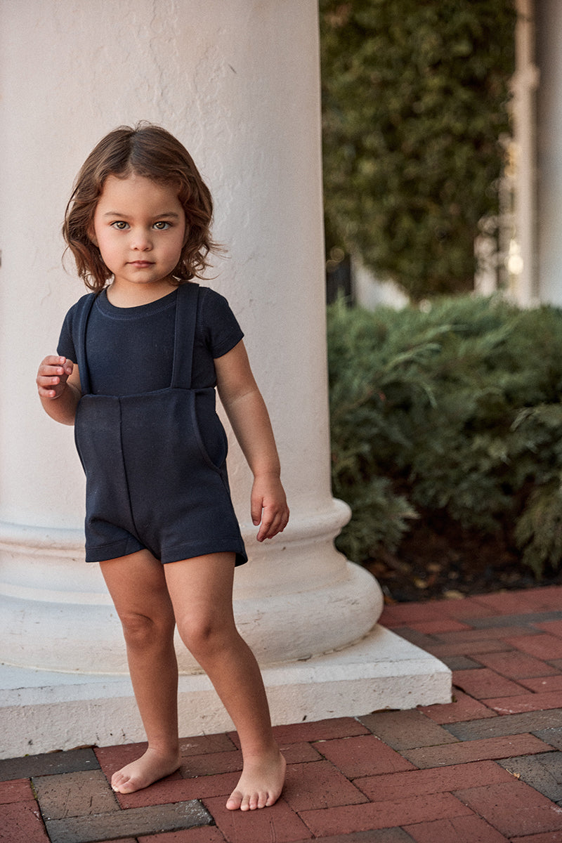 Child wearing a navy blue romper standing on a brick path with a white column and greenery in the background.