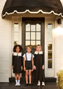 Three children standing on a porch with a black awning and door.