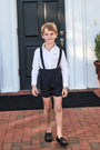 Young boy in suspenders standing in front of a house with a black door.