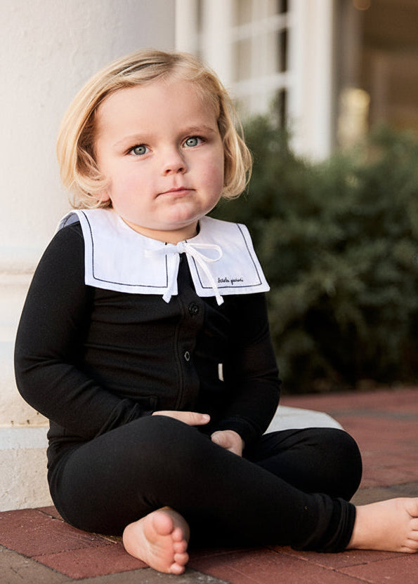 Child wearing a black outfit with a white collar sitting on a brick surface.