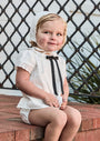 Child wearing a white outfit with a black bow tie and bonnet, sitting on a brick ledge.
