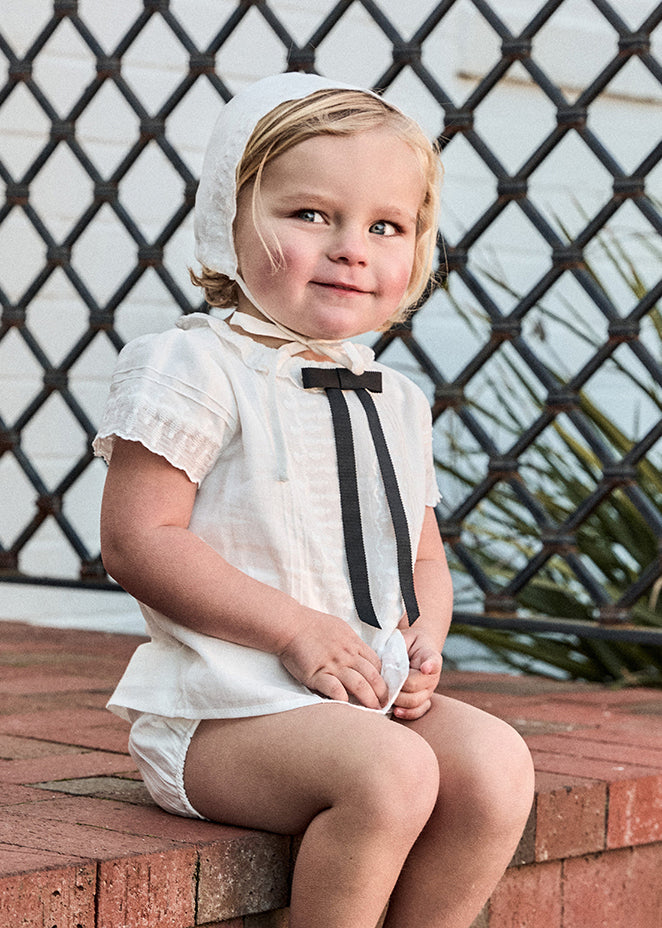 Child wearing a white outfit with a black bow tie and bonnet, sitting on a brick ledge.
