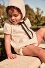 Child wearing a white bonnet and outfit with a black bow sitting outdoors.