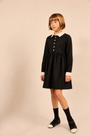 A young girl with short brown hair stands in a studio wearing a black long-sleeve dress with a white pointed collar and white button placket, paired with black socks and black-and-white shoes, against a plain beige background.