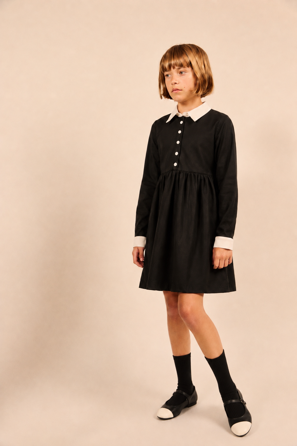 A young girl with short brown hair stands in a studio wearing a black long-sleeve dress with a white pointed collar and white button placket, paired with black socks and black-and-white shoes, against a plain beige background.