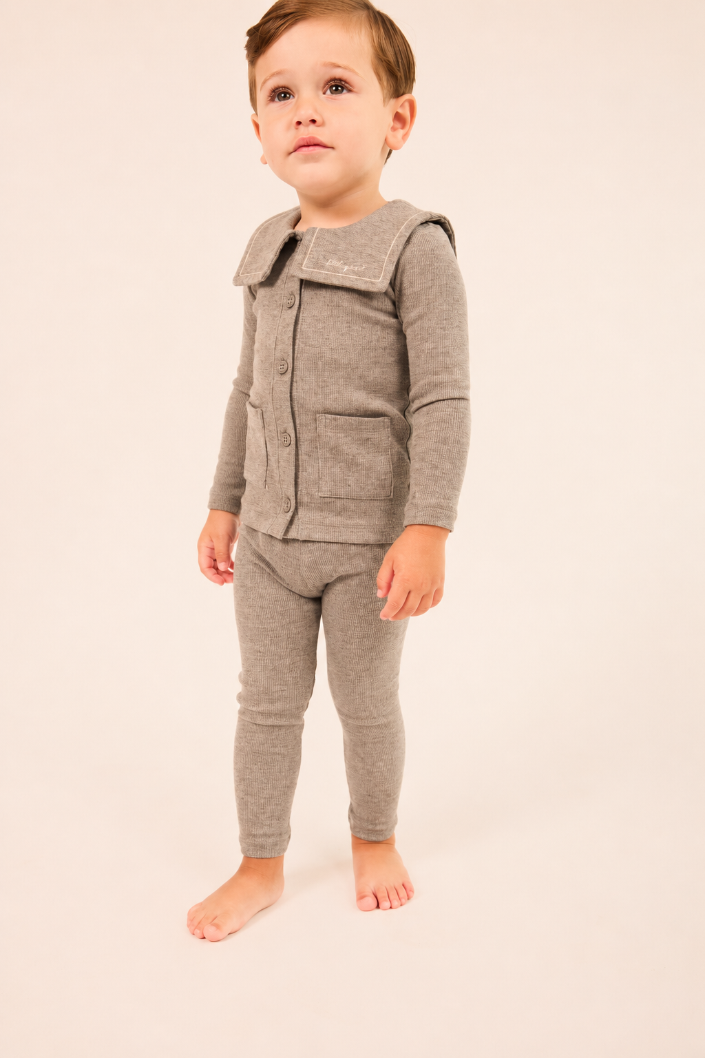 A toddler standing barefoot against a plain light beige studio background, wearing a matching heather gray knit outfit with a long-sleeve button-front top and leggings.