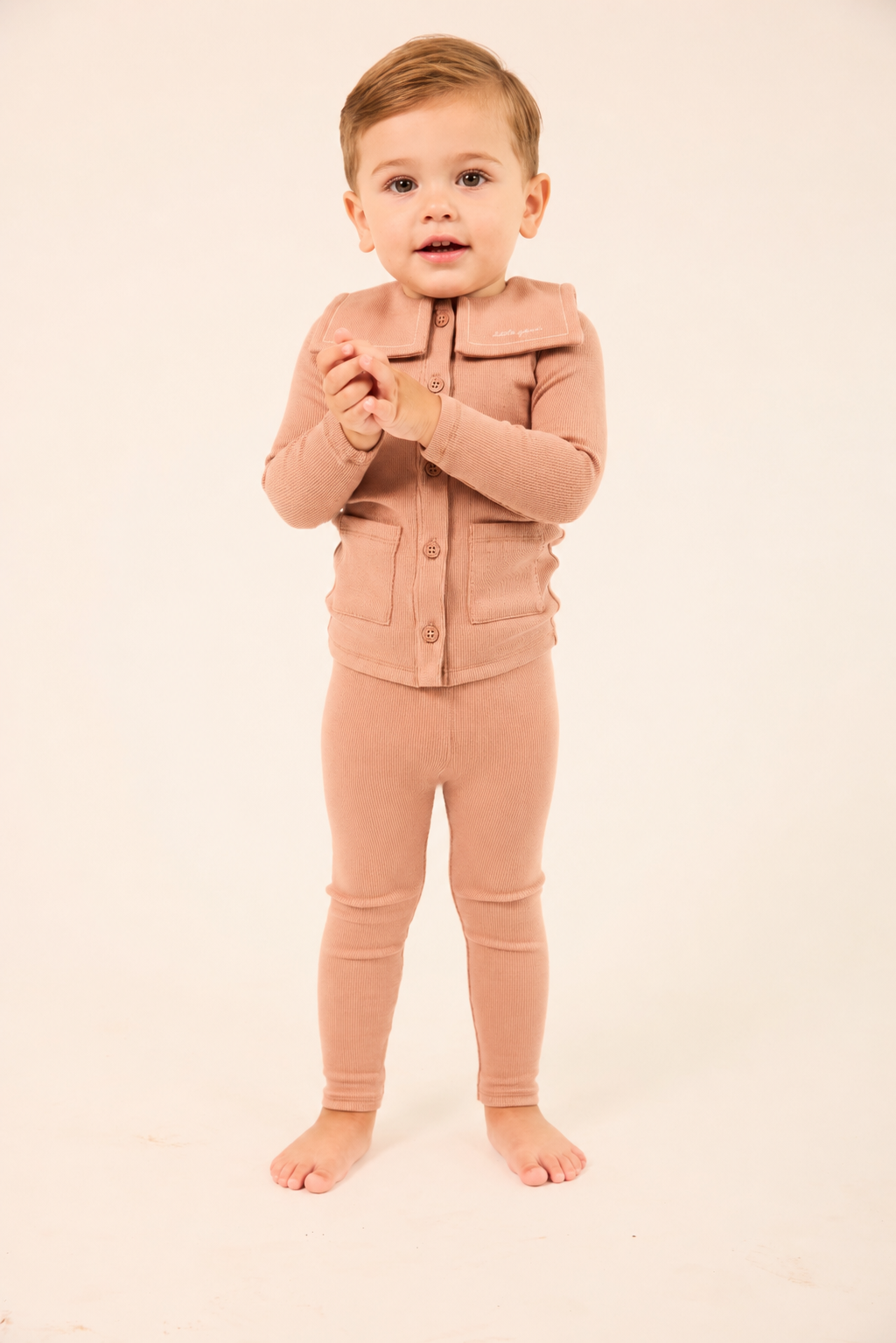 A toddler standing barefoot against a plain cream studio background, wearing a matching dusty rose cardigan and leggings set.