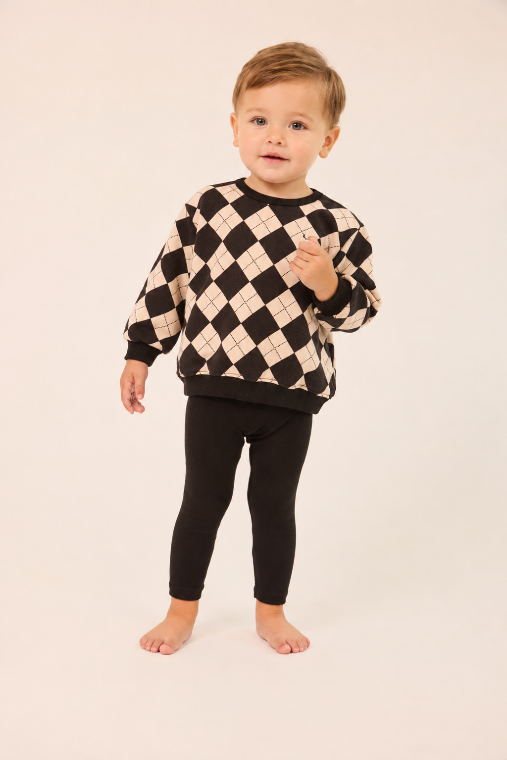 A toddler standing barefoot against a plain light beige studio background, wearing a black and cream argyle sweater with black leggings.