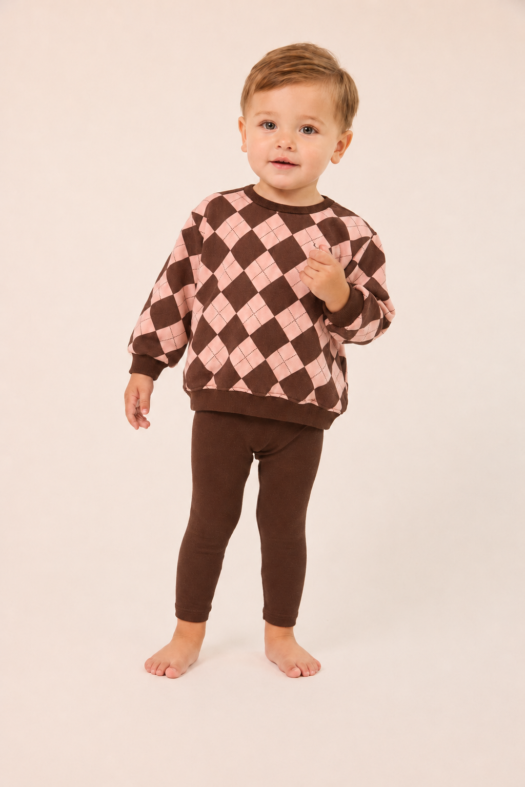 A toddler standing barefoot against a plain light beige studio background, wearing a long-sleeve argyle sweater in brown and light pink with matching brown leggings.