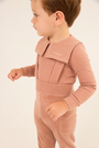A toddler wearing a dusty rose pink ribbed corduroy jumpsuit with a wide collar and shoulder straps, shown in profile against a plain light background.