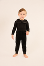 A toddler standing barefoot against a plain light beige studio background, wearing a black long-sleeve jumpsuit with a structured collar and elastic waist.